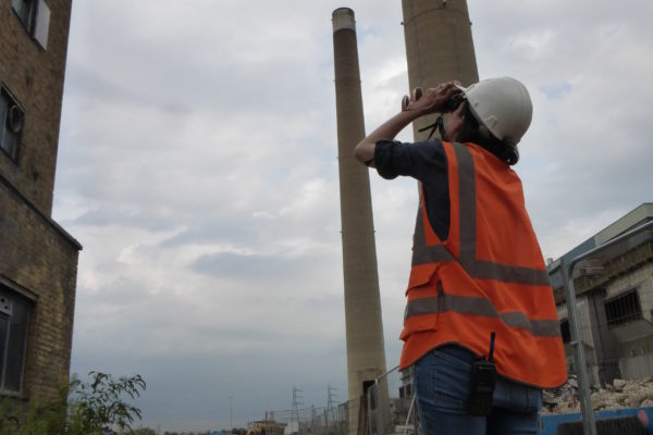 Rebecca Read looking upwards through binoculars in an industrial site with tall chimneys.