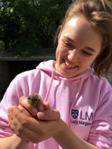 A young woman in a pink hooded top holds a bird out to the camera whilst smiling.